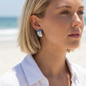 May include: Silver-toned seashell-shaped stud earrings. The earrings have a smooth, polished finish and a curved design, reflecting light. The earrings are worn on the ear of a person with blonde hair, against a blurred beach background.