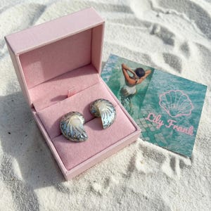 Könnte beinhalten: Ein Paar muschelförmige Ohrringe mit irisierendem Glanz und silberfarbenen Filigranmustern, präsentiert in einer offenen hellrosa Schmuckschatulle. Daneben liegt eine rechteckige Karte mit einem Foto einer Person im Wasser und dem Schriftzug Lily Frank.