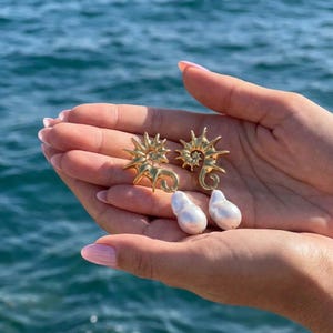 May include: Gold-colored seahorse earrings with white pearl drops. The earrings have a detailed, spiky design and are displayed against a backdrop of blue water. The pearls are teardrop-shaped.