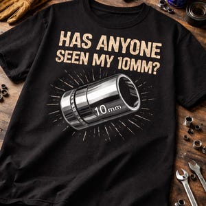 以下が含まれることがあります： 「HAS ANYONE SEEN MY 10MM？」のテキストと10mmソケットのグラフィックがプリントされた黒いTシャツ。ソケットはシルバーで、「10mm」の測定値が印刷されています。デザインはサンバースト効果があります。