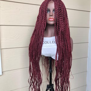 May include: A long, burgundy braided wig with a lace front. The wig is styled in a straight, loose braid pattern. The wig is displayed on a mannequin head.