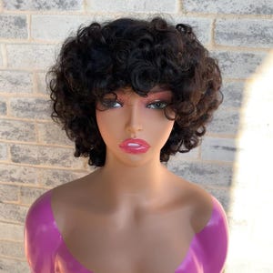May include: A black, curly wig styled with a short, layered cut. The wig is displayed on a mannequin head with a pink bust. The background is a light gray brick wall.