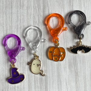 Può includere: Quattro chiusure a moschettone in plastica viola, trasparente, arancione e nera, ciascuna attaccata a un ciondolo in metallo. I ciondoli raffigurano un cappello da strega viola con stelle dorate, un fantasma bianco, una zucca arancione e un pipistrello nero con dettagli dorati.