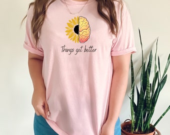 May include: Pink t-shirt featuring a graphic of a sunflower and a brain with the text "Things get better".