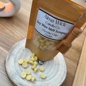 May include: A brown paper bag with a white label that reads "Alma Wick Candle Co. Soy Wax Melt Scoopies Chocolate Chip Cookie". The bag is filled with small, round, white wax melts. A wooden scoop is next to the bag.