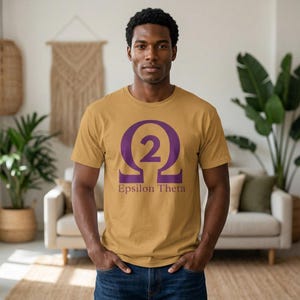 May include: A mustard yellow short-sleeved t-shirt featuring a large purple Omega symbol with the number 2 centered inside. Below the symbol, the text Epsilon Theta is printed in a matching purple font across the chest of the garment.