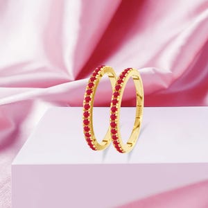 Könnte beinhalten: Zwei goldfarbene Ringe mit einer Reihe kleiner, roter Edelsteine. Die Ringe werden auf einer weißen Oberfläche vor einem rosa Hintergrund präsentiert. Die Edelsteine sind gleichmäßig um den Umfang jedes Rings angeordnet.