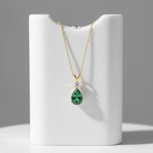 Puede incluir: Un collar de oro con un colgante de piedra preciosa verde esmeralda en forma de pera. El colgante está acentuado con una pequeña piedra preciosa redonda y transparente. El collar se exhibe sobre un soporte blanco.