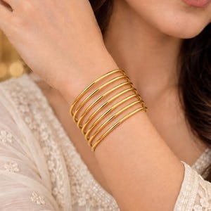 Op de afbeelding: Een set van zeven dunne, gepolijste goudkleurige armbanden die samen om de pols worden gedragen. Elke armband heeft een glad, afgerond profiel met een reflecterende metallic afwerking, wat zorgt voor een gestapelde look rond de arm.