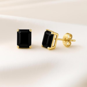 May include: A pair of gold stud earrings with rectangular, black gemstones. The earrings have a classic design with a post and butterfly backing. The gemstones are set in a gold bezel setting.
