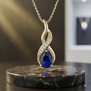 May include: A gold pendant necklace with a pear-shaped, deep blue gemstone. The pendant features an infinity-style design with small, clear gemstones and hangs from a delicate gold chain. The necklace is displayed on a marble pedestal.