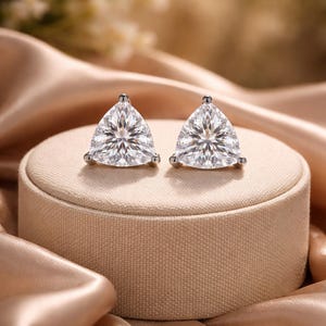 May include: A pair of stud earrings featuring triangular, clear gemstones. The earrings are set in silver-coloured metal and are displayed on a beige, round jewellery box, against a backdrop of a satin fabric.