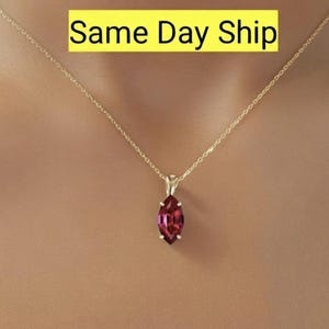 Op de afbeelding: Een gouden ketting met een hanger met een marquise-geslepen, dieprode edelsteen. De ketting wordt gepresenteerd tegen een huidkleurige achtergrond. Tekst in een gele doos luidt "Same Day Ship".