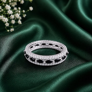 May include: A silver-toned band ring featuring a central row of marquise-cut black stones flanked by two rows of small, round, clear white stones. The ring has a metallic finish and a symmetrical design with alternating stone shapes and colors.