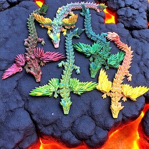 Könnte beinhalten: Eine Sammlung farbenfroher, beweglicher Drachenfiguren. Jeder Drache hat ein einzigartiges Farbschema, darunter Rosa, Grün, Gelb und Blau, mit detaillierten Flügel- und Körperdesigns. Die Drachen sind auf einer dunklen, strukturierten Oberfläche mit einem feurigen, orangefarbenen Glühen angeordnet.