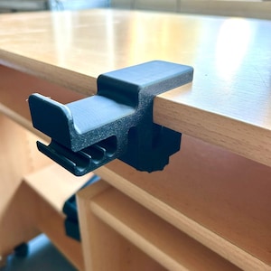Könnte beinhalten: Eine schwarze Kunststoffklemme, die an der Kante eines Holztisches befestigt ist. Die Klemme hat eine rechteckige Oberseite und einen gebogenen, geschlitzten Boden. Der Tisch ist hellbraun, und der Hintergrund ist unscharf.