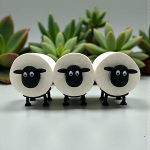 Könnte beinhalten: Drei weiße Schaf-Figuren mit schwarzen Gesichtern, Ohren und Beinen sind in einer Reihe angeordnet. Die Schafe haben eine runde Körperform und stehen vor einem Hintergrund aus grünen Sukkulenten. Die Figuren sind klein und dekorativ.