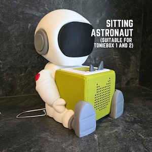Puede incluir: Una figura de astronauta blanca con visera negra y detalles grises, sosteniendo un Toniebox verde lima. El texto en la imagen dice "SITTING ASTRONAUT (SUITABLE FOR TONIEBOX 1 AND 2)". El astronauta está sentado.
