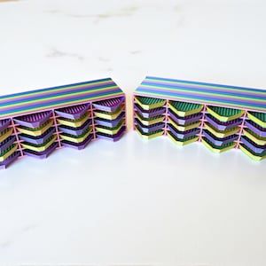 Könnte beinhalten: Zwei farbenfrohe, geometrische Aufbewahrungsboxen. Jede hat einen rosa Rahmen mit mehreren abgewinkelten Fächern in Lila, Gelb und Grün. Die Oberseiten weisen horizontale Streifen in Regenbogenfarben auf. Auf einer weißen Oberfläche.