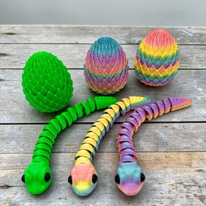 May include: A collection of 3D-printed toys, including three articulated snakes in green, yellow, and rainbow colors, and three dragon eggs in green and rainbow hues. The toys are arranged on a wooden surface.