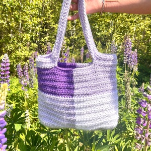 May include: A crocheted tote bag with a purple and white ombre design. The bag has two straps and is made with a textured stitch.