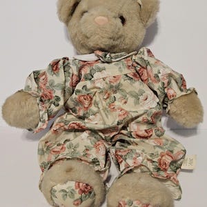 May include: A beige teddy bear wearing a floral print onesie with pink and green roses. The bear has a pink nose and brown eyes.