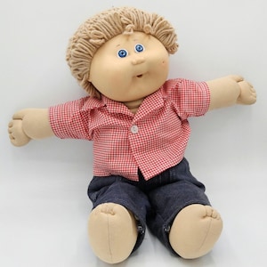 May include: A soft-sculpture doll with light skin, bright blue eyes, and tan yarn hair. The doll wears a red and white gingham button-down shirt with a single white button and dark blue denim pants. The doll has a rounded face and limbs.