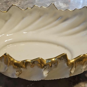May include: White ceramic serving bowl with a gold rim and a shell-like design. The bowl has a raised handle on one side.