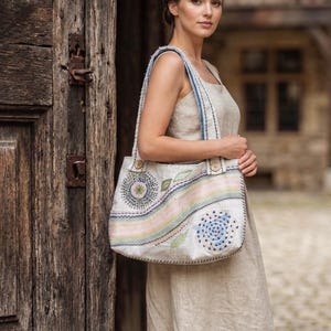 Może przedstawiać: Beżowa torba typu tote z haftowanymi kwiatowymi i geometrycznymi wzorami w pastelowych kolorach. Torba ma jasnoniebieski pasek i drewniane guziki. Torba jest noszona przez osobę ubraną w beżową sukienkę.