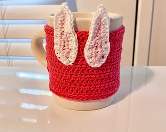 Op de afbeelding: Een witte keramische mok met een roze gehaakte cozy met twee witte gehaakte konijnenoren.