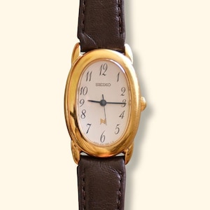 May include: A Seiko wristwatch with an oval gold-tone case and a dark brown leather strap. The white dial features black Arabic numerals, black hour and minute hands, and a gold-tone logo symbol above the six o'clock position.