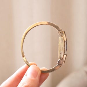 Może przedstawiać: Zegarek na bransolecie w stylu vintage w kolorze złotym. Zegarek ma prostokątną tarczę i zawiasową konstrukcję bransoletki. Bransoletka jest trzymana otwarta przez dłoń, prezentując profil boczny zegarka. Zegarek ma klasyczną, elegancką estetykę.