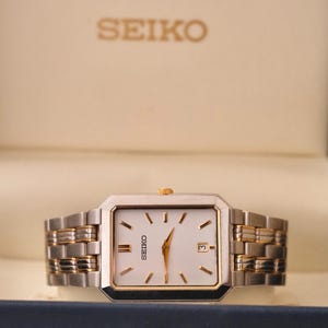 May include: A rectangular Seiko watch with a white face, gold hands, and a date display at the 3 o'clock position. The watch has a two-tone silver and gold band. The Seiko logo is visible on the watch face and the box.