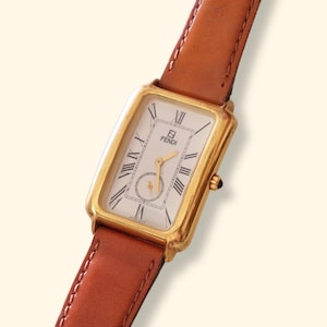 Può includere: Orologio Fendi rettangolare color oro con quadrante bianco, numeri romani e piccolo quadrante dei secondi. Cinturino in pelle marrone. Il logo Fendi è visibile sul quadrante.