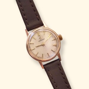 May include: A vintage Omega wristwatch featuring a round gold-tone case with a cream-colored dial, gold-tone hour markers, and hands. The watch is attached to a dark brown leather strap with visible stitching and a small gold-tone crown on the side.