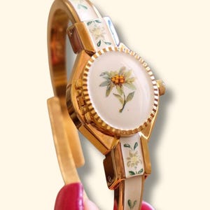 Op de afbeelding: Een goudkleurig armbandhorloge met een witte geëmailleerde wijzerplaat met een centrale afbeelding van een edelweissbloem. De scharnierende armband is versierd met bijpassende witte emaille panelen en kleine bloemmotieven, met een geribbelde goudkleurige rand.