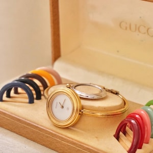Puede incluir: Un reloj de pulsera rígida en tono dorado con esfera circular blanca y el logotipo de Gucci, presentado en una caja color canela con múltiples biseles intercambiables de colores. El reloj cuenta con una banda dorada fina y corona.
