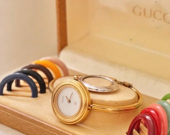 May include: A gold-tone bangle watch with a white circular face and the Gucci logo, presented in a tan box with multiple interchangeable colored bezels. The watch features a thin gold band and a small crown, accompanied by a silver-tone bezel.