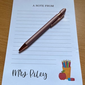 May include: A white notepad with lined paper and the text "A Note From Mrs. Riley" printed on it. A rose gold pen is lying on the notepad. The notepad has a colourful illustration of a pencil holder with pencils, an apple, and an eraser.