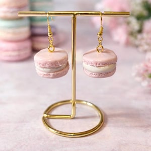 Könnte beinhalten: Rosa Macaron-Ohrringe mit goldfarbenen Haken und einem goldfarbenen Präsentationsständer. Die Ohrringe sind wie Macarons mit einer weißen Füllung geformt. Weitere Macarons sind im Hintergrund gestapelt.