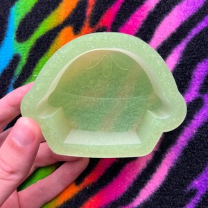May include: A light green, glittery, silicone mold shaped like a cartoon character with a large head and a small body. The mold is designed to create a resin or polymer clay figurine.