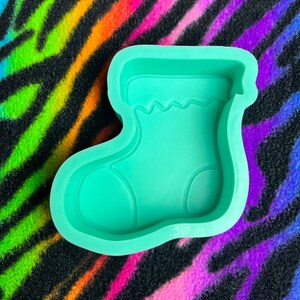 May include: A green silicone mold shaped like a Christmas stocking. The mold has a detailed design with a ruffled top and a rounded toe.