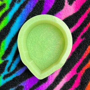 May include: A light green silicone mold shaped like an alien head. The mold has a textured surface and a raised alien face design.