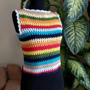 Puede incluir: Un top de crochet sin mangas con un patrón de rayas arcoíris. El top tiene un ribete de crochet blanco alrededor del escote.