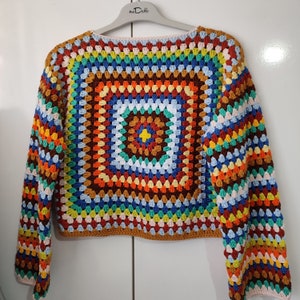 Può includere: Un maglione all'uncinetto colorato con maniche lunghe e un motivo quadrato in tonalità di rosso, arancione, giallo, verde, blu e marrone.