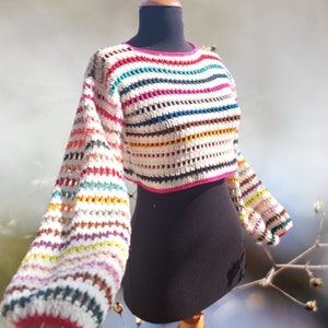 May include: A white crochet top with rainbow stripes and long sleeves. The top is cropped and has a pink trim around the neckline.