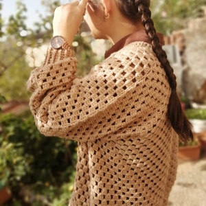 Può includere: Un cardigan all'uncinetto beige con un motivo quadrato. Il cardigan ha maniche lunghe e viene indossato sopra una camicia marrone con colletto. Un orologio in oro rosa è visibile al polso. La persona indossa occhiali e ha una lunga treccia.