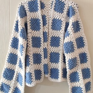 Könnte beinhalten: Eine weiße und blaue gehäkelte Strickjacke mit quadratischem Muster. Die Strickjacke hat lange Ärmel und eine lockere Passform.