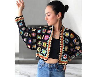 Puede incluir: Una chaqueta de punto de ganchillo negra con cuadrados de granny coloridos. La chaqueta tiene una longitud recortada y mangas largas. La chaqueta está hecha con una variedad de colores, incluyendo rojo, naranja, amarillo, verde, azul y morado.