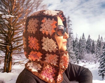 May include: A brown crocheted hooded cowl with pink and white accents. The cowl is worn by a person in a snowy landscape.